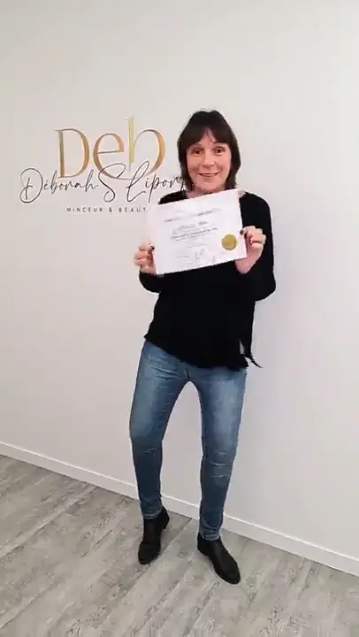 Une stagiaire en jean et haut foncé tient son attestation à deux mains devant le mur de marque.
