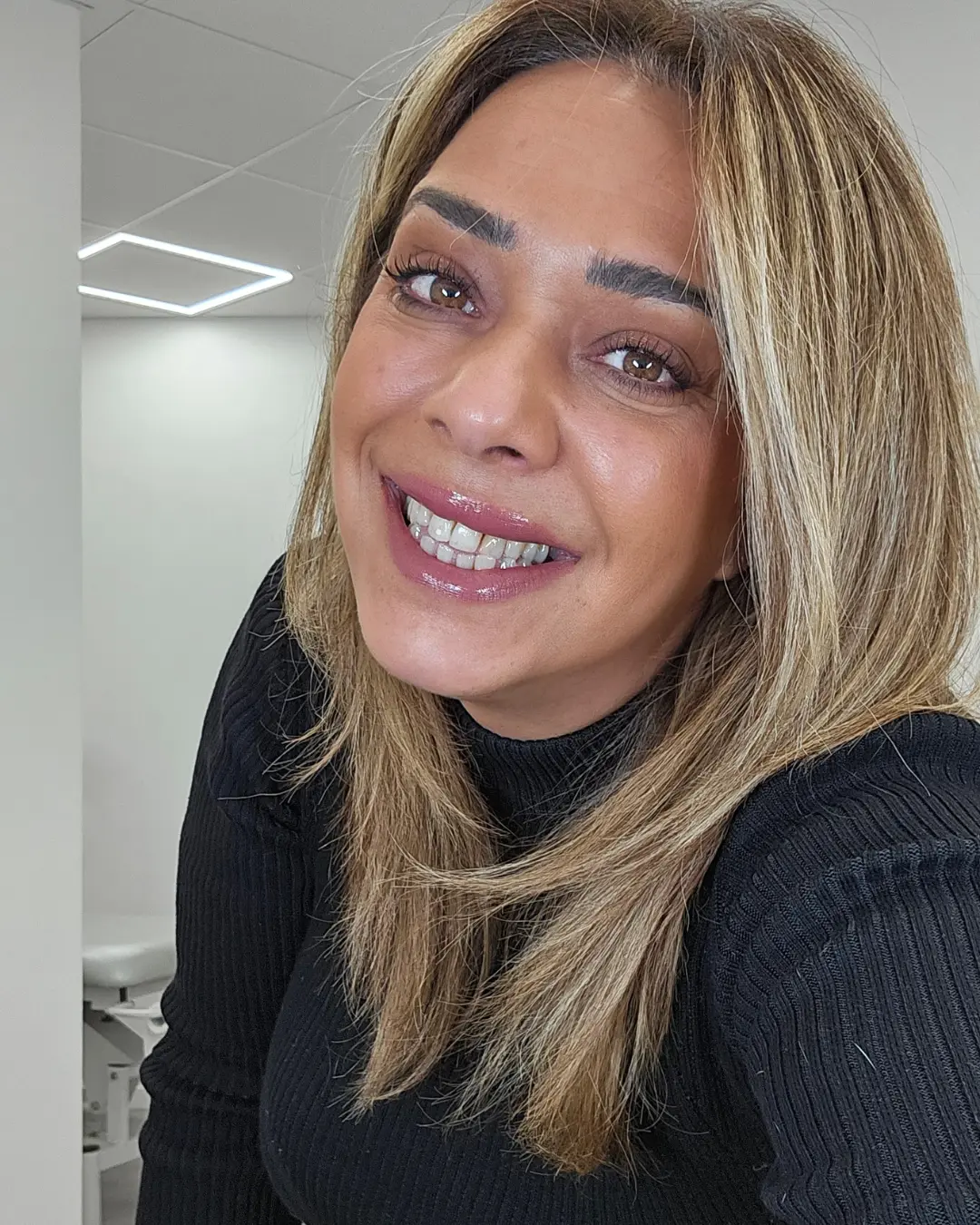 La praticienne, cheveux blonds et haut noir, sourit face à l’objectif dans la cabine, sous le plafonnier.