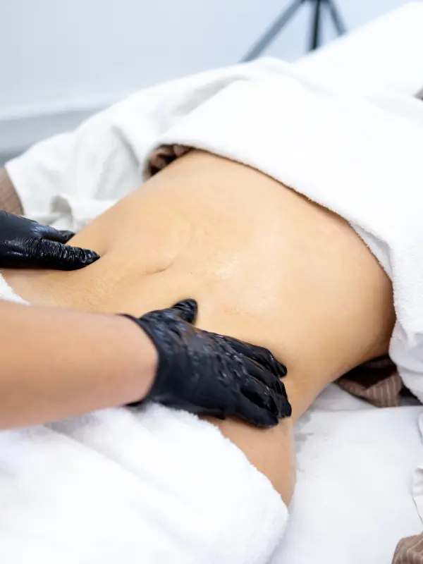 Deux mains gantées de noir travaillent le ventre d’une cliente, une serviette blanche repliée au-dessus.